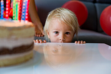 Beautiful two years old toddler boy in blue shirt, celebrating his birthday, blowing candles on homemade baked cake, indoors