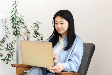 Freelance concept. Asian young woman sits in an armchair with a laptop working at home.