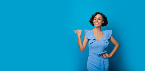 Close-up photo of a happy beautiful young woman in a blue dress, who is pointing with help of her finger and isolated on blue background
