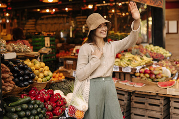 Friendly young caucasian brunette girl waving looking to side standing at street market. Pretty woman wears casual clothes and shopping bag. Healthy food, natural products concept