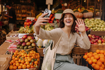 Joyful young caucasian woman waving at phone screen in fruit market row. Brunette wears casual clothes, hat and shopping bag. Technology concept