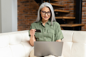 Calm senior businesswoman in glasses sitting at the sofa using laptop and credit card for online payment from home, typing number to enter internet banking account, ordering food
