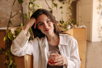 Nice young caucasian woman looks at camera, holds glass with drink in room. Brunette wears top and shirt on casual day. People sincere emotions lifestyle concept.