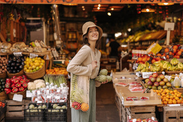 Smiling young caucasian woman carries shopping bag with vegetables shopping at markets during day. Brunette in casual clothes is smiling looking at camera. Lifestyle concept