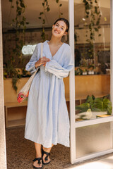Naklejka premium Full length pretty young caucasian woman with shopping bag looks at camera standing in doorway. Brunette wears blue sundress in summer. Lifestyle, different emotions, leisure concept.