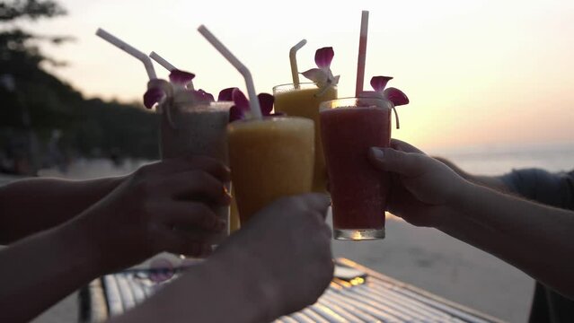 Friends sit in a beach cafe overlooking the sea and amazing sunset clink tasty cocktails in their hands.