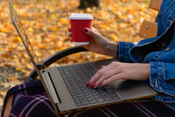 Close-up to the hand young woman holding a cup of coffee while using a laptop in a autumn park.