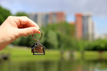 Keychain in shape of a house in female hand on background of summer lake and high-rise residential...