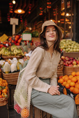 Modern young caucasian woman looking at camera sitting at fruit and vegetable market. Brunette wears blouse, pants, hat and string bag. Concept natural products