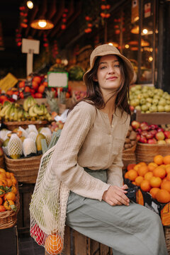 Modern young caucasian woman looking at camera sitting at fruit and vegetable market. Brunette wears blouse, pants, hat and string bag. Concept natural products