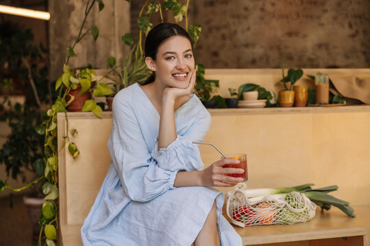 Smiling young caucasian woman looking at camera enjoying cool summer drink indoors. Brunette with collected hair wears casual sundress. Good mood concept