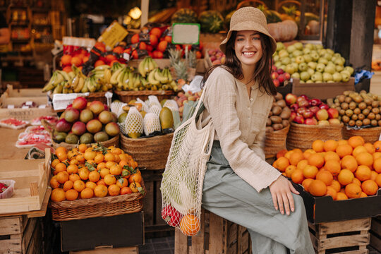 Smiling young caucasian girl looking at camera spending time near fruit stand in summer. Brunette wears casual clothes, hat and shopping bag. Concept of natural products