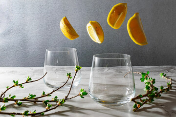 Transparent thin glasses made of glass, wide shape, lemon slices fly nearby and a branch from a currant bush lies, gray background, ease of serving