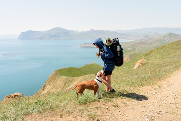 Young beautiful woman in hood travel with backpack on mountains road and playing with her dog