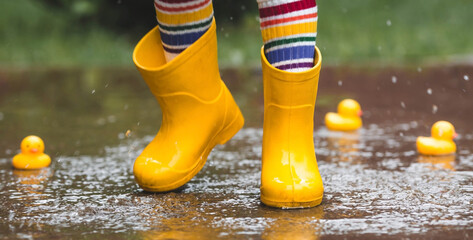 A small child in rainbow-colored socks and yellow rubber boots jumps through puddles and plays with...