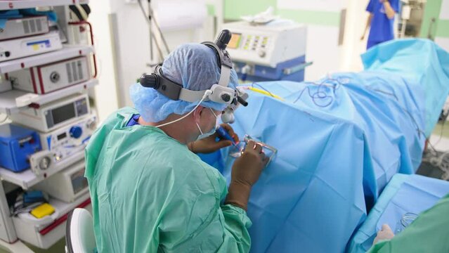 Doctor in protective clothes and device glasses sits over the patient using instruments. View of the surgeon from his back.