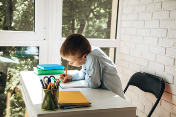 A schoolboy writes numbers in a notebook at a white desk. Back to school concept