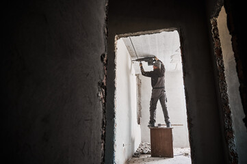 Full length of man builder standing on wooden table and using electric drill while making hole in wall. View through doorway of male worker drilling wall in room under renovation.