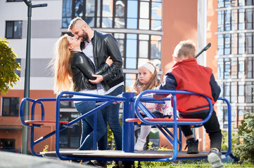 Obraz premium Happy parents hanging out with kids, while children swinging at playground. Front view of two adorable kids spinning on merry-go-round, while parents hugging behind outdoor. Concept of family.