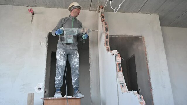 Full length of man in workwear drilling wall with hammer drill. Male worker using drill breaker while destroying wall in apartment under renovation. Demolition work and home renovation concept.