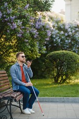 Young blind man with smartphone sitting on bench in park in city, calling.