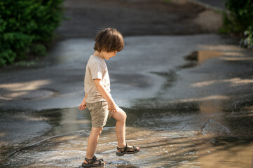 a cute three-year-old Asian boy with long black hair happily stomps through puddles. Happy children walking outside after the rain on a hot sunny summer day