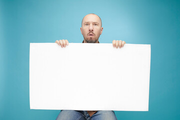  A Bald man with emotion face holding  white board in his hands to fill your text. Isolated on blue background.