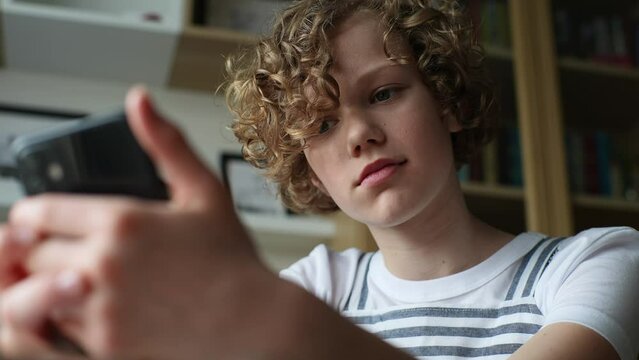 Close-up low-angle view of cheerful curly little 10s girl holding smartphone, watching funny video, distance chatting with friends from home. Adorable kid studying online in mobile app using phone.