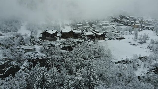 Cozy winter village in the mountains of French Alps covered in clouds and snow with wooden chalet houses drone zoom out through fog.