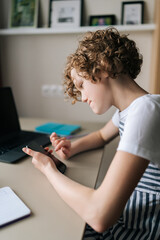 Fototapeta premium Vertical shot of focused adorable preteen school girl e-learning, doing homework projects remotely, preparing for test, tutoring online from home using laptop and smartphone, sitting at desk by window