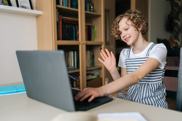 Smiling little curly girl talking happily with friends through video conferencing on laptop computer sitting at table in modern bedroom. Positive adorable kid using laptop for e-learning online.