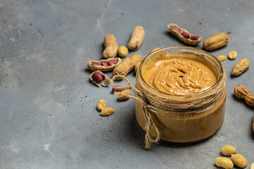 Peanut butter or paste in an open jar and peanuts in the peel scattered on a gray background, banner, menu, recipe place for text, top view
