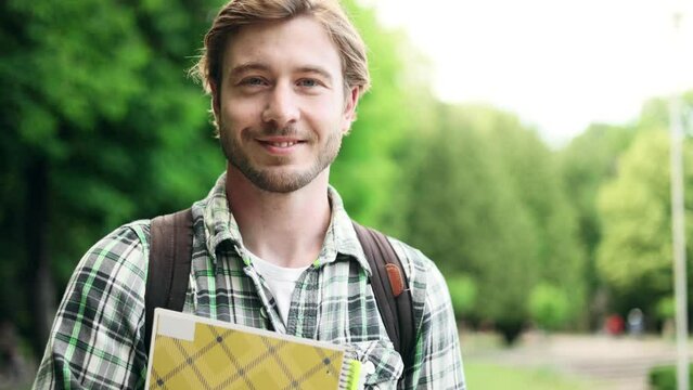 Portrait of student guy standing with books near the park and looking at the camera. Portrait of attractive blond man.
