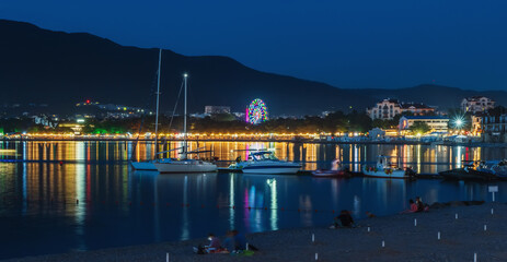 Fototapeta na wymiar Night photo of sea bay with yachts and boats and reflection of resort town illumination in sea water against backdrop of mountains and blue sky.
