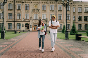 Two international stylish students walk near the campus and smile. Friendly African American guy and Caucasian girl walk together near the university