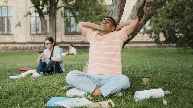 General scene of a campus life. Focus on african American student with glasses putting down his books and relaxing with his eyes closed