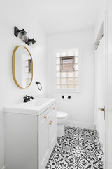 A bathroom with a white vanity cabinet, mosaic black and white tiled floor, and a gold mirror.