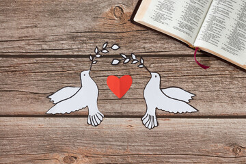 Two paper doves carry branches, an open Holy Bible Book, and a red heart on a wooden background....