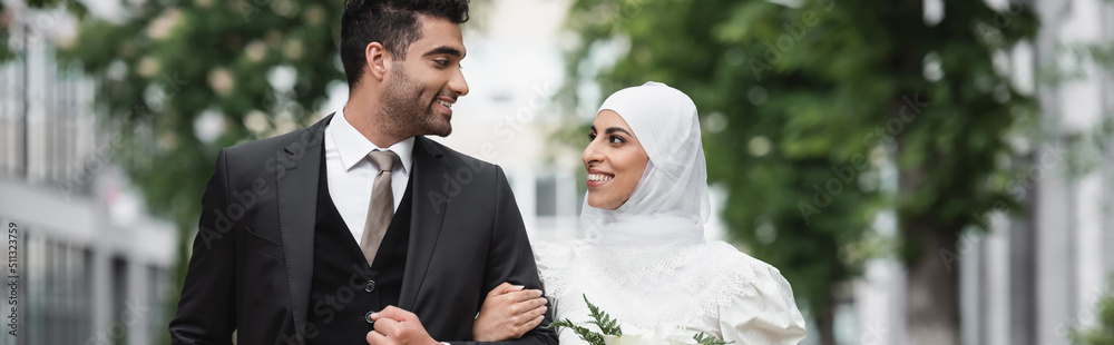 Wall mural happy muslim bride in wedding hijab and white dress holding bouquet near groom outside, banner.