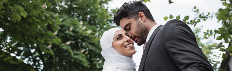 groom in suit hugging cheerful muslim bride in white hijab, banner.