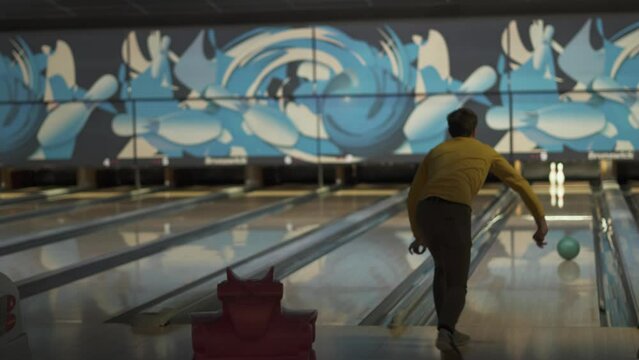 Thin man throws dark green ball into bowling alley, watches throw. Ball rolls along edge of track past pins. Optimistic bowler smiles, waves hand. Mental health, accepting mistakes with positivity.