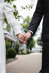 cropped view of newlyweds in wedding dress and formal wear holding hands outdoors.