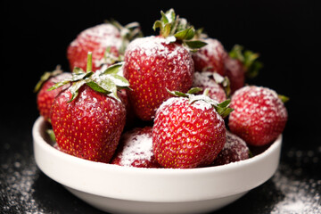 Fototapeta na wymiar Strawberries in a plate with powdered sugar on a black background, side view close-up.