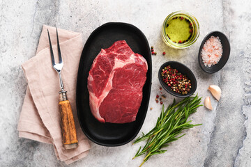 Beef steak. Fresh raw beef rib eye steak with fork rosemary, salt and pepper on a grill pan on grey stone background. Top view. Mockup for design idea.