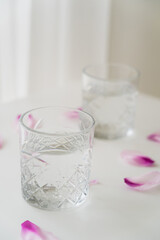close up view of faceted glass with clean water near floral petals on blurred grey background.