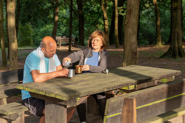 Fototapeta premium Senior couple sitting in a picnic table in the forest drkinking water from bottles in metal camping cups