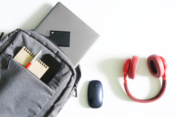 Gray urban backpack or bag with laptop, external ssd drive, mobile phone, notepad and pen on a white table next to mouse and red headphones. Tools and gadgets of a student or freelancer.