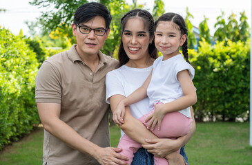 Asian mother holds her six-year-old daughter standing together with her father, a warm and happy family in the backyard.