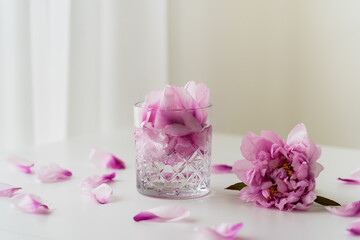 Obraz na płótnie Canvas faceted glass with tonic and petals near pink peony on white tabletop and grey background.