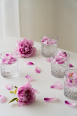 Obraz na płótnie Canvas crystal glasses with tonic and floral petals near pink peonies on white tabletop and grey background.